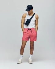 A man wearing a black cap, white tank top, Psycho Bunny MENS SAMOS HYDROCHROMIC PRINTED SWIM TRUNK - B6W453F200 in pink, white socks, white sneakers, and a black waterproof crossbody bag stands with hands in pockets looking down.
