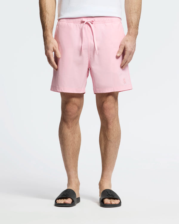 A person in light pink Psycho Bunny MENS SAMOS HYDROCHROMIC PRINTED SWIM TRUNKS, featuring innovative hydrochromic technology, stands against a plain white background wearing a white shirt and black slide sandals.