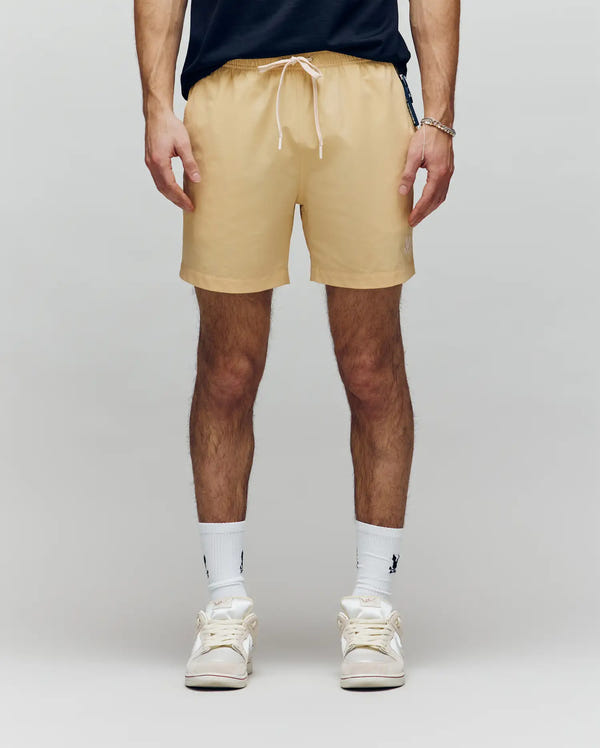 A man stands facing the camera against a plain light gray background, wearing Psycho Bunny MENS SAMOS HYDROCHROMIC PRINTED SWIM TRUNK (B6W453F200), a dark T-shirt, white socks, and white sneakers. His hands are relaxed by his sides.