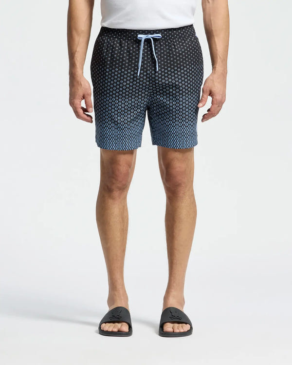 A person wears a white shirt, Psycho Bunny's MENS BENDIGO ALL OVER PRINTED SWIM TRUNK (B6W619F200) in gradient blue and black, and black slide sandals with a logo. The shorts have no-chafe netting and a drawstring waist. The background is plain white.