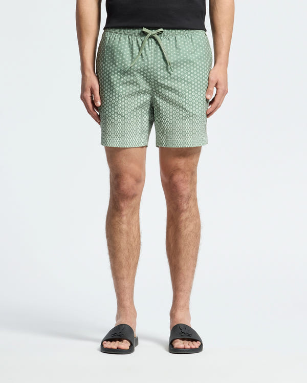 A person wears Psycho Bunny's MENS BENDIGO PRINTED SWIM TRUNK (B6W619F200) with an elasticized waist and drawstring, along with black slide sandals. Made of quick-dry fabric, these light green patterned shorts provide both style and function. Upper body is cropped against a white background.