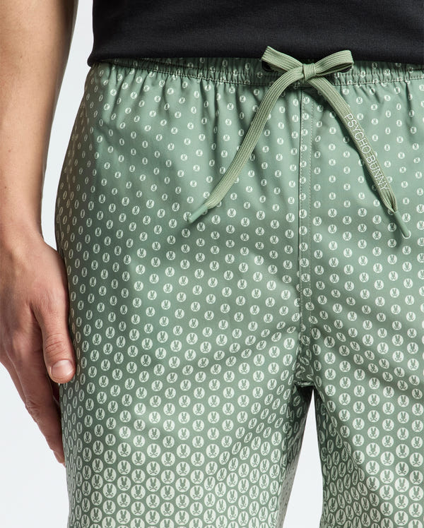 Someone is wearing the MENS BENDIGO PRINTED SWIM TRUNK by Psycho Bunny, which features a light green color with a small white pattern and an elasticized drawstring waist. The slightly glossy texture of the trunks complements their black top as their hand rests casually on the side.