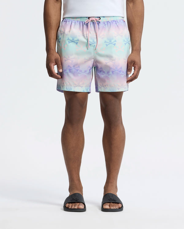 Dressed in Psycho Bunny's MENS FORSTER BUNNY PRINTED SWIM TRUNK (B6W621F200), a person stands relaxed, exuding beach-bum comfort. Black slip-on sandals complement the pastel swim shorts’ star pattern, set against a white background, capturing an ocean view vibe.