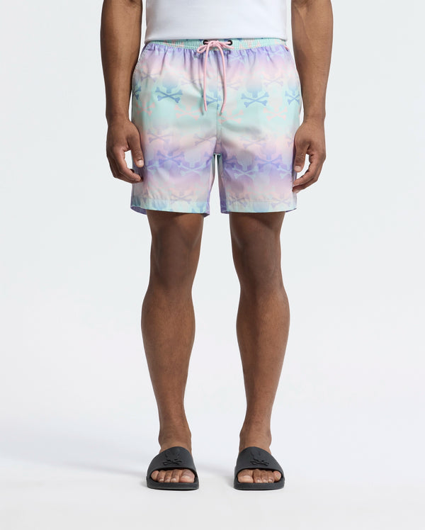 Dressed in Psycho Bunny's MENS FORSTER BUNNY PRINTED SWIM TRUNK (B6W621F200), a person stands relaxed, exuding beach-bum comfort. Black slip-on sandals complement the pastel swim shorts’ star pattern, set against a white background, capturing an ocean view vibe.
