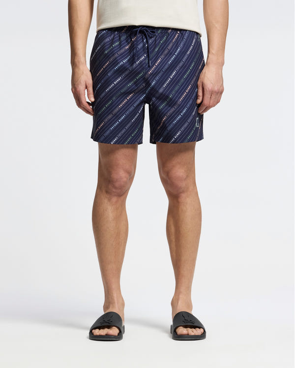 A person in a white shirt and Psycho Bunny Men's Renmark PB Printed Swim Trunk - B6W622F200 stands against a light background. These quick-dry trunks feature diagonal lines and small text, complemented by black slides. The stylish shorts with a logo-patterned print are the highlight of the look.