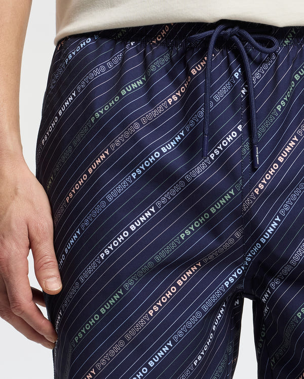 Close-up of a person wearing Psycho Bunny's Men's Renmark PB Printed Swim Trunk, featuring a vibrant multi-color logo pattern. A drawstring waist ensures comfort, while a partially visible hand adds intrigue to the stylish navy design.