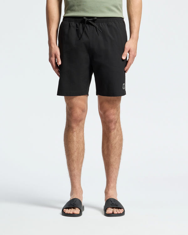 A person is wearing Psycho Bunny's black MENS SAMOS 7 HYDROCHROMIC SWIM TRUNK with a drawstring and black slide sandals featuring a textured design, paired with a green top. The sunsoaker style highlights the lower body against a plain background.