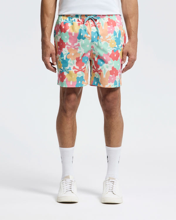 A man adorned in Psycho Bunny's Mens Lemmon All Over Print Swim Trunk (B6W658F200) is seen wearing colorful floral shorts in pink, yellow, blue, and peach hues. Paired with a white T-shirt, white socks, and sneakers, he stands against a plain backdrop.
