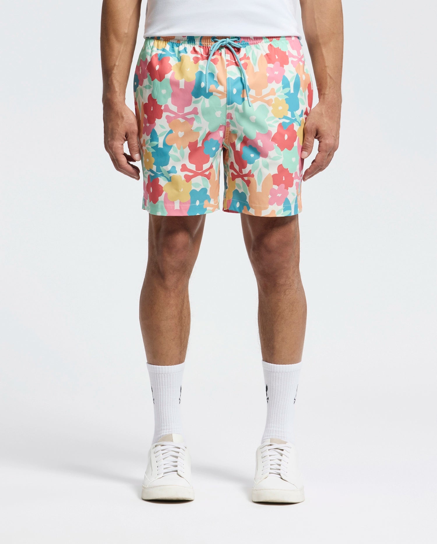 A man adorned in Psycho Bunny's Mens Lemmon All Over Print Swim Trunk (B6W658F200) is seen wearing colorful floral shorts in pink, yellow, blue, and peach hues. Paired with a white T-shirt, white socks, and sneakers, he stands against a plain backdrop.