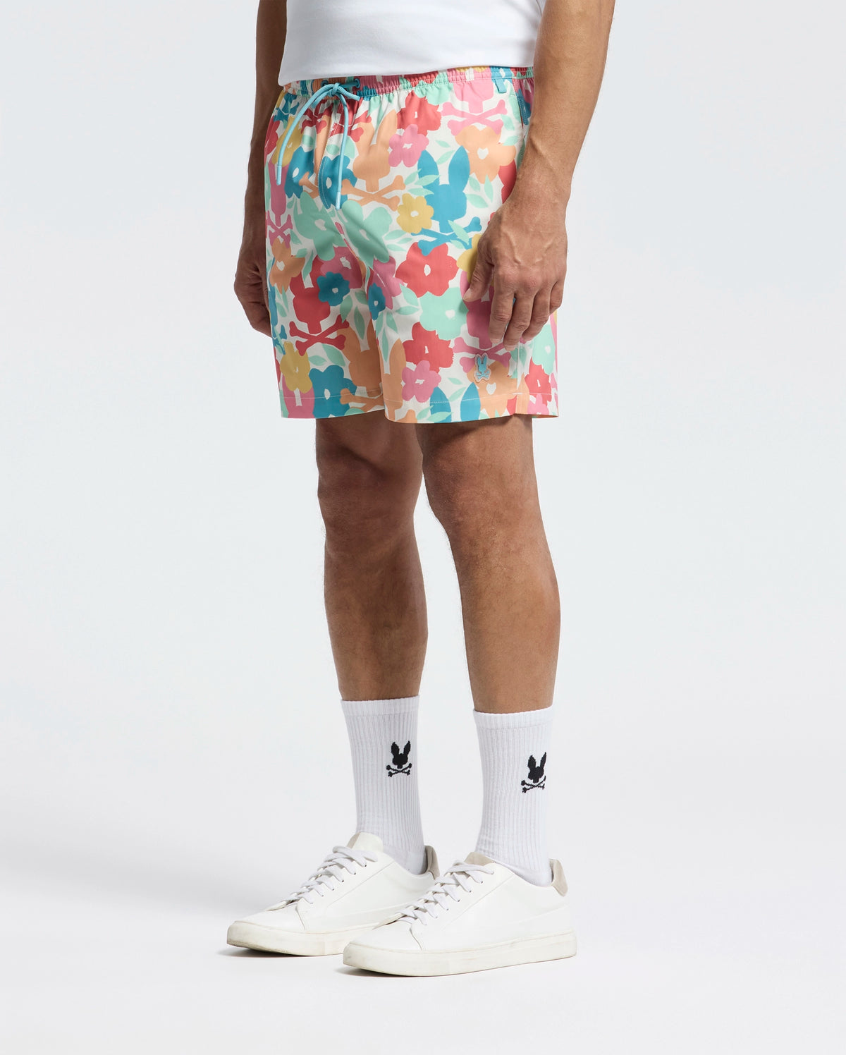 A person stands in a white shirt, Psycho Bunny's MENS LEMMON ALL OVER PRINT SWIM TRUNK featuring red, pink, blue, and yellow floral patterns. Complementing the vibrant trunks are comfy white socks with a black logo and clean white sneakers against a plain white backdrop.