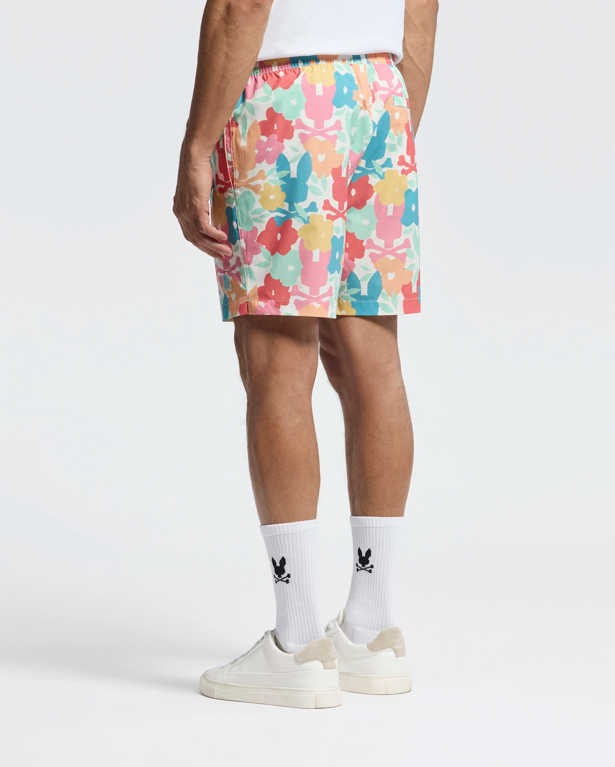 A person is sporting the vibrant Psycho Bunny MENS LEMMON ALL OVER PRINT SWIM TRUNK, featuring an abstract design in pink, orange, blue, and green on a white background, paired with a white T-shirt, black logo white socks, and white sneakers.