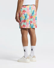 A person is sporting the vibrant Psycho Bunny MENS LEMMON ALL OVER PRINT SWIM TRUNK, featuring an abstract design in pink, orange, blue, and green on a white background, paired with a white T-shirt, black logo white socks, and white sneakers.