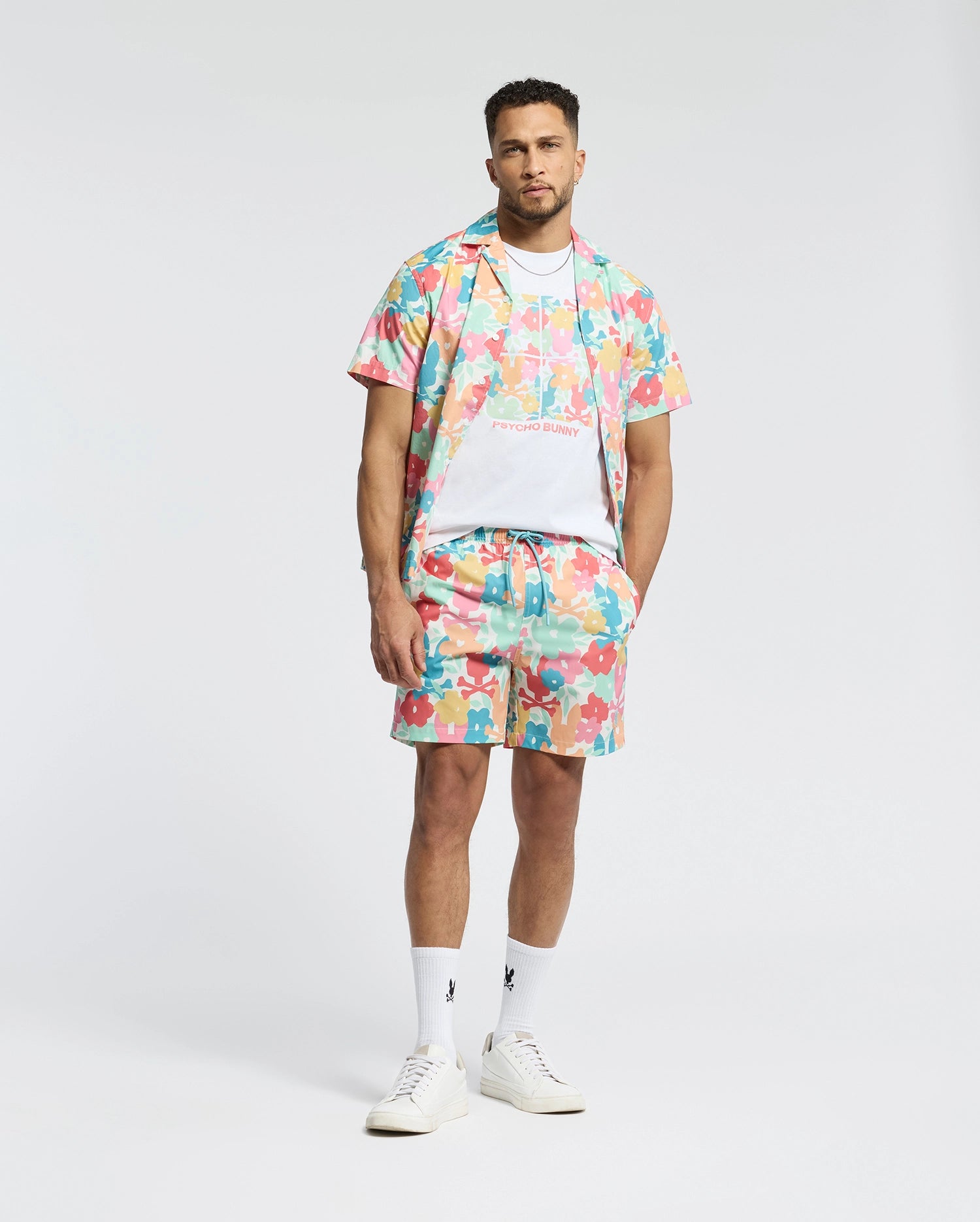 A man in Psycho Bunny's vibrant MENS LEMMON ALL OVER PRINT SWIM TRUNK and matching shirt stands against a plain background. His style is effortlessly showcased with white sneakers, socks, and a T-shirt underneath.