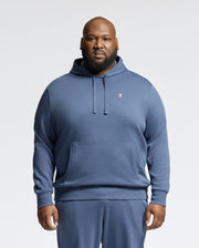 A man faces forward in a blue Psycho Bunny MENS BIG AND TALL LEVY ESSENTIAL FLEECE HOODIE (B9H859D200) and matching sweatpants. Made from soft 100% cotton French terry, the hoodie has a small white embroidered logo on the chest. The background is plain white.