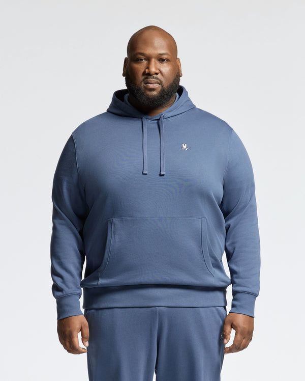 A man faces forward in a blue Psycho Bunny MENS BIG AND TALL LEVY ESSENTIAL FLEECE HOODIE (B9H859D200) and matching sweatpants. Made from soft 100% cotton French terry, the hoodie has a small white embroidered logo on the chest. The background is plain white.