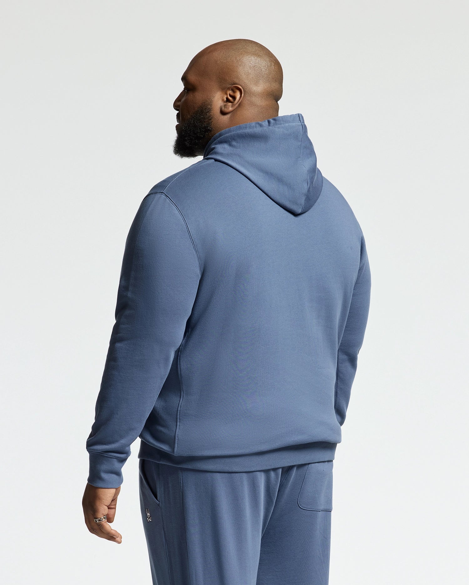 A bearded man stands with his back to the camera, wearing a blue Psycho Bunny MENS BIG AND TALL LEVY ESSENTIAL FLEECE HOODIE (B9H859D200) and matching sweatpants, set against a plain white background.