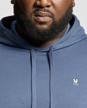 A close-up of a man wearing the Psycho Bunny MENS BIG AND TALL LEVY ESSENTIAL FLEECE HOODIE, featuring a small embroidered bunny and crossbones logo on the chest, showing only his lower face, beard, and neck.