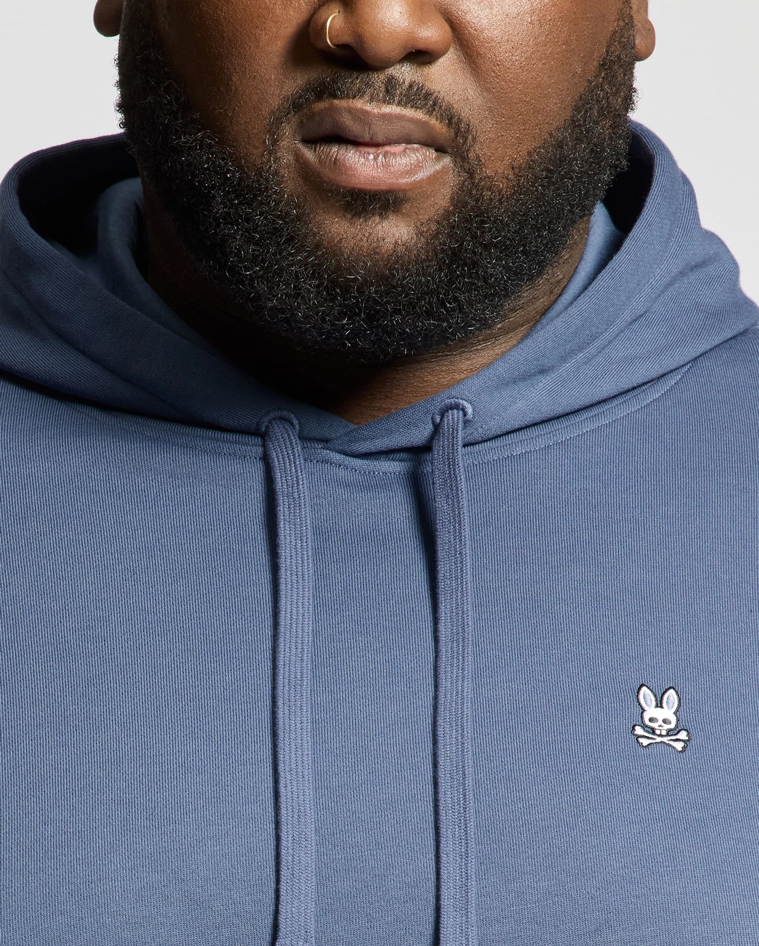 A close-up of a man wearing the Psycho Bunny MENS BIG AND TALL LEVY ESSENTIAL FLEECE HOODIE, featuring a small embroidered bunny and crossbones logo on the chest, showing only his lower face, beard, and neck.