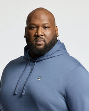 A bald, bearded man wears a blue Psycho Bunny MENS BIG AND TALL LEVY ESSENTIAL FLEECE HOODIE (B9H859D200), made from 100% cotton French terry with a small bunny logo on the chest, standing against a plain light background.