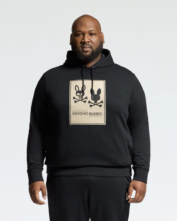 A man confidently stands against a plain white background, wearing a Psycho Bunny MENS BIG AND TALL MONROE TWILL PATCH FLEECE HOODIE. This black fleece features two cartoon bunnies with crossbones and an applique patch of the iconic "Psycho Bunny" logo. Crafted from cozy French terry, it's stylishly unique.