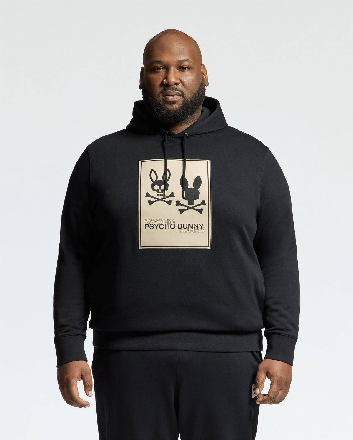 A man confidently stands against a plain white background, wearing a Psycho Bunny MENS BIG AND TALL MONROE TWILL PATCH FLEECE HOODIE. This black fleece features two cartoon bunnies with crossbones and an applique patch of the iconic "Psycho Bunny" logo. Crafted from cozy French terry, it's stylishly unique.