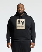 A man confidently stands against a plain white background, wearing a Psycho Bunny MENS BIG AND TALL MONROE TWILL PATCH FLEECE HOODIE. This black fleece features two cartoon bunnies with crossbones and an applique patch of the iconic "Psycho Bunny" logo. Crafted from cozy French terry, it's stylishly unique.