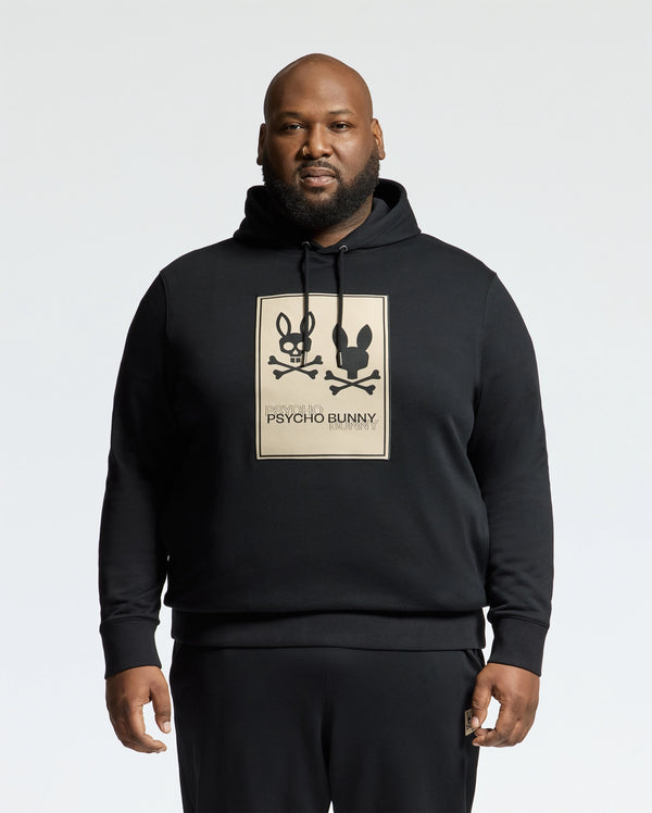 A man confidently stands against a plain white background, wearing a Psycho Bunny MENS BIG AND TALL MONROE TWILL PATCH FLEECE HOODIE. This black fleece features two cartoon bunnies with crossbones and an applique patch of the iconic "Psycho Bunny" logo. Crafted from cozy French terry, it's stylishly unique.