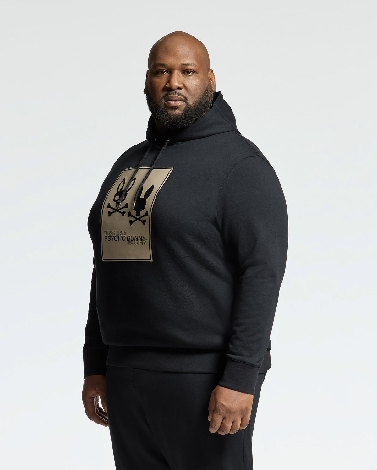 A man confidently wears a Psycho Bunny men's big and tall Monroe black twill patch fleece hoodie, featuring a beige graphic and logo on the front. The simple light gray backdrop highlights his pose and attire.