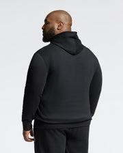 A bearded, shaved-head person faces away from the camera wearing a Psycho Bunny black Monroe Twill Patch Fleece Hoodie and matching black pants, against a plain light background.