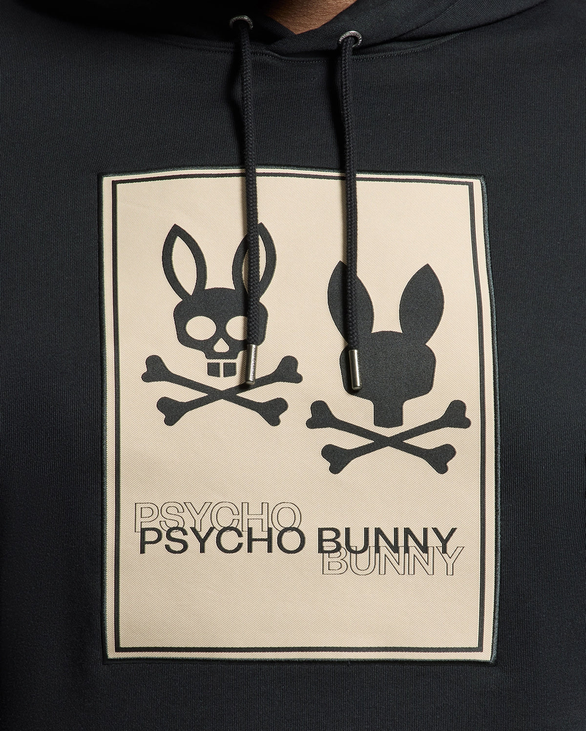 The Psycho Bunny Men's Big and Tall Monroe Twill Patch Fleece Hoodie features a black design with two bunny skull and crossbones graphics, complemented by the "PSYCHO PSYCHO BUNNY BUNNY" phrase in alternating shades, finished with a signature patch applique.
