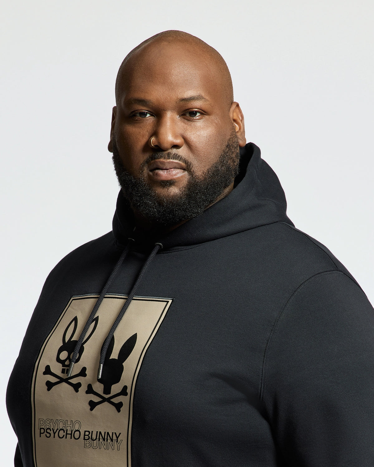 A bearded person wears the Psycho Bunny Monroe Hoodie, crafted from soft French terry. The black hoodie features a large square print with the "Psycho Bunny" logo against a plain, light background.