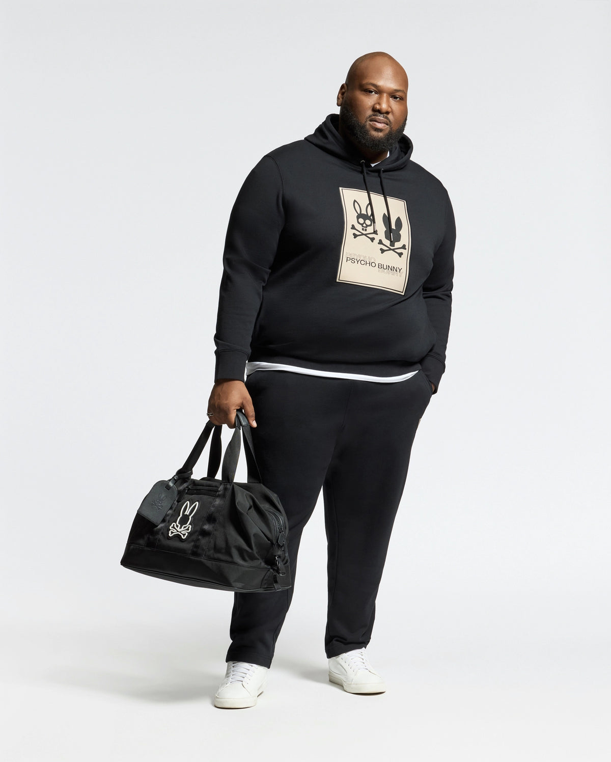A man stands against a plain background wearing Psycho Bunny's black mens big and tall Monroe twill patch fleece hoodie with a cartoon graphic, black pants, holding a black duffel bag in his left hand, and white sneakers, creating a casual yet stylish look.