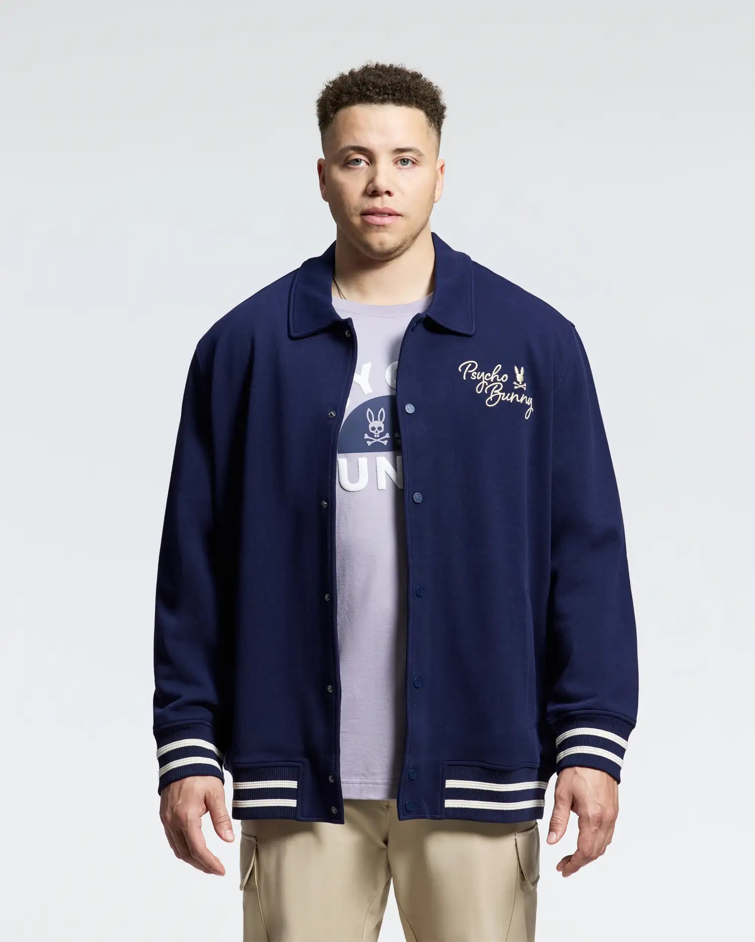 A man wearing a navy blue Men's Big and Tall Desert Coach Bomber Jacket (B9J428F200) with "Psycho Bunny" on the chest, layered over a lavender T-shirt showing "BUN". The cotton blend outerwear complements his short curly hair, against a plain background.