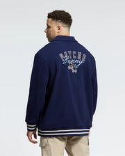 A man models the Psycho Bunny Men's Big and Tall Desert Coach Bomber Jacket B9J428F200 in navy, featuring white and grey striped cuffs and hem, with "Psycho Bunny" text and a bunny design on the back. Made from a cotton blend, he stands relaxed, gazing sideways.