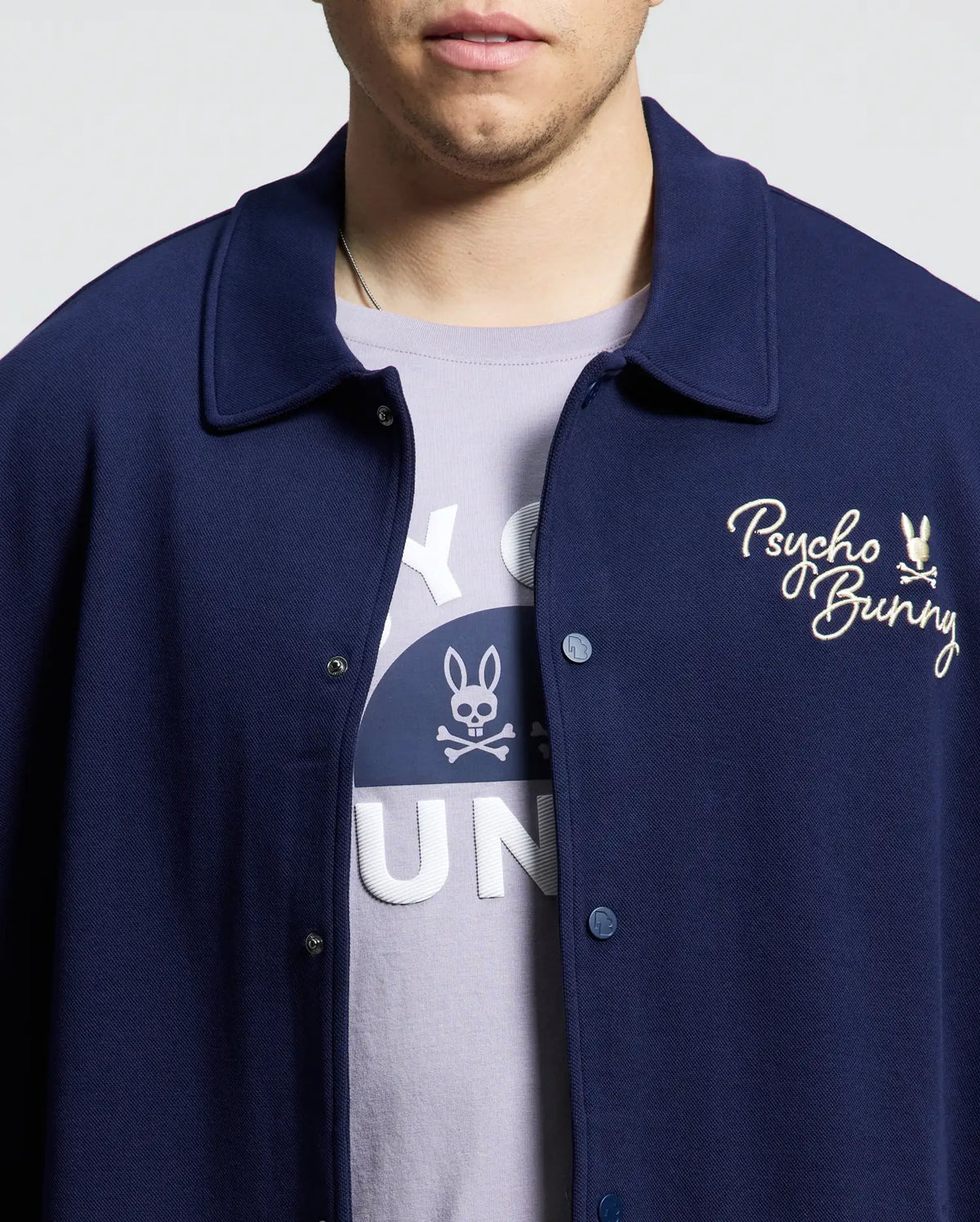 A person is wearing the MENS BIG AND TALL DESERT COACH BOMBER JACKET - B9J428F200 by Psycho Bunny in navy blue with "Psycho Bunny" embroidery. Underneath, they have a light purple, soft cotton blend T-shirt featuring a bunny graphic. Their face is partially visible.