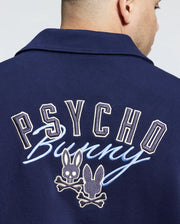 A person wearing the navy Psycho Bunny MENS BIG AND TALL DESERT COACH BOMBER JACKET (B9J428F200) with an embroidered back design featuring "Psycho Bunny" text, two bunny heads—one with eyes and a skull motif—and crossed bones, is facing away.