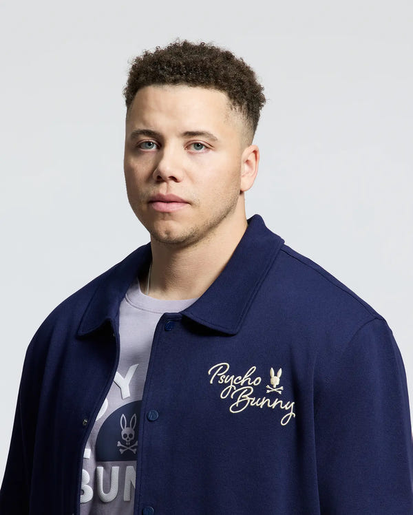 A person with short curly hair wears a navy blue MENS BIG AND TALL DESERT COACH BOMBER JACKET - B9J428F200 over a patterned shirt. This cotton blend jacket, featuring "Psycho Bunny" embroidery on the chest, contrasts with the plain light gray background.