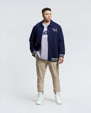 A person in a Psycho Bunny MENS BIG AND TALL DESERT COACH BOMBER JACKET - B9J428F200 stands against a plain background, wearing a light shirt, beige cargo pants, and white high-tops. They have short hair and hands in their pockets.