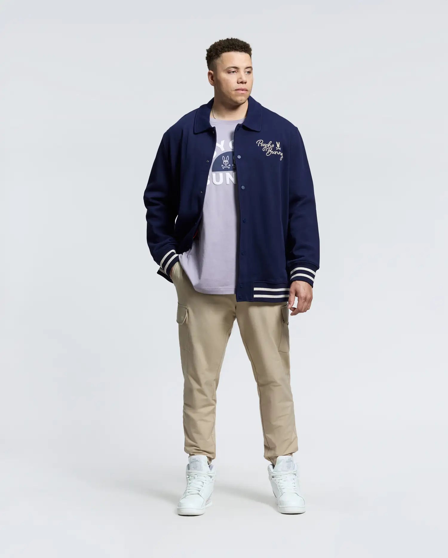 A person in a Psycho Bunny MENS BIG AND TALL DESERT COACH BOMBER JACKET - B9J428F200 stands against a plain background, wearing a light shirt, beige cargo pants, and white high-tops. They have short hair and hands in their pockets.