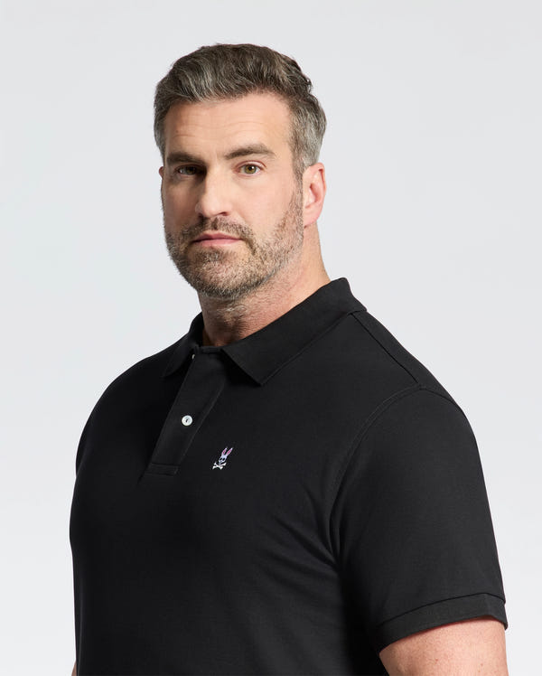 A man with short hair and a beard wears the Psycho Bunny Mens Big and Tall Classic Pique Polo Shirt (B9K001ARPC) featuring a small embroidered logo, standing against a plain light background.