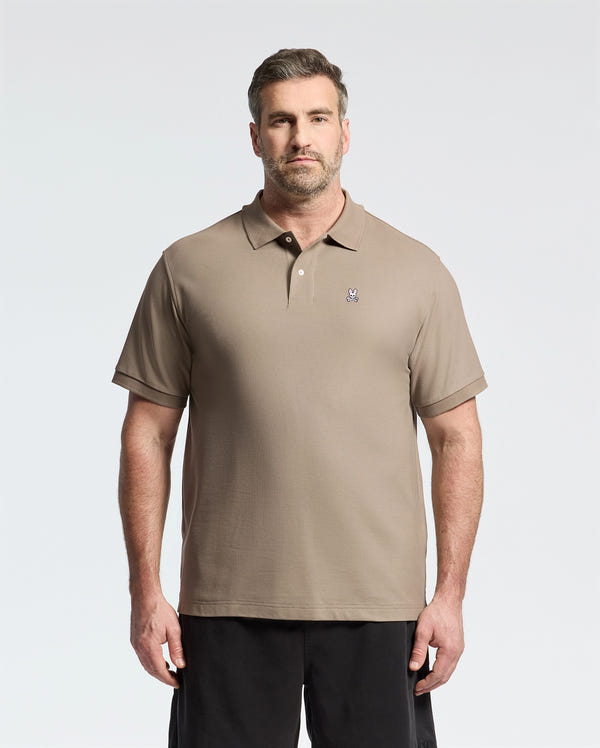 A man in a Psycho Bunny MENS BIG AND TALL CLASSIC PIQUE POLO SHIRT - B9K001CRPC, light brown with an embroidered logo on the left chest, stands against a plain background. Made from soft pima cotton, the shirt complements his short hair and beard and pairs stylishly with dark pants.