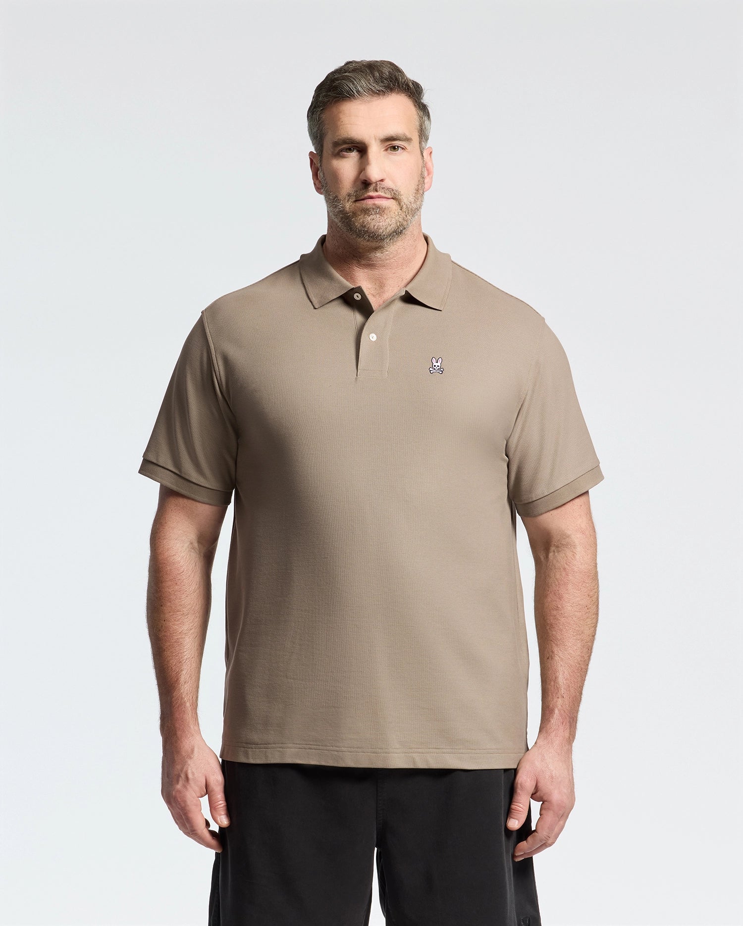 A man in a Psycho Bunny MENS BIG AND TALL CLASSIC PIQUE POLO SHIRT - B9K001CRPC, light brown with an embroidered logo on the left chest, stands against a plain background. Made from soft pima cotton, the shirt complements his short hair and beard and pairs stylishly with dark pants.