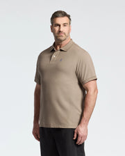 A man with short hair and a beard is wearing the Psycho Bunny MENS BIG AND TALL CLASSIC PIQUE POLO SHIRT in taupe and dark pants, standing against a light gray background. He gazes slightly left with a neutral expression.