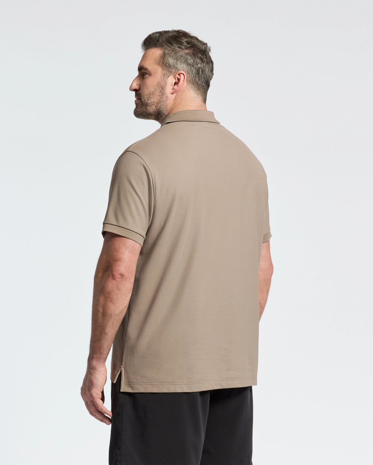 A man with short hair stands sideways, facing left, wearing the Psycho Bunny Mens Big and Tall Classic Pique Polo Shirt in beige pima cotton and black shorts against a plain white background.