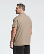 A man with short hair stands sideways, facing left, wearing the Psycho Bunny Mens Big and Tall Classic Pique Polo Shirt in beige pima cotton and black shorts against a plain white background.