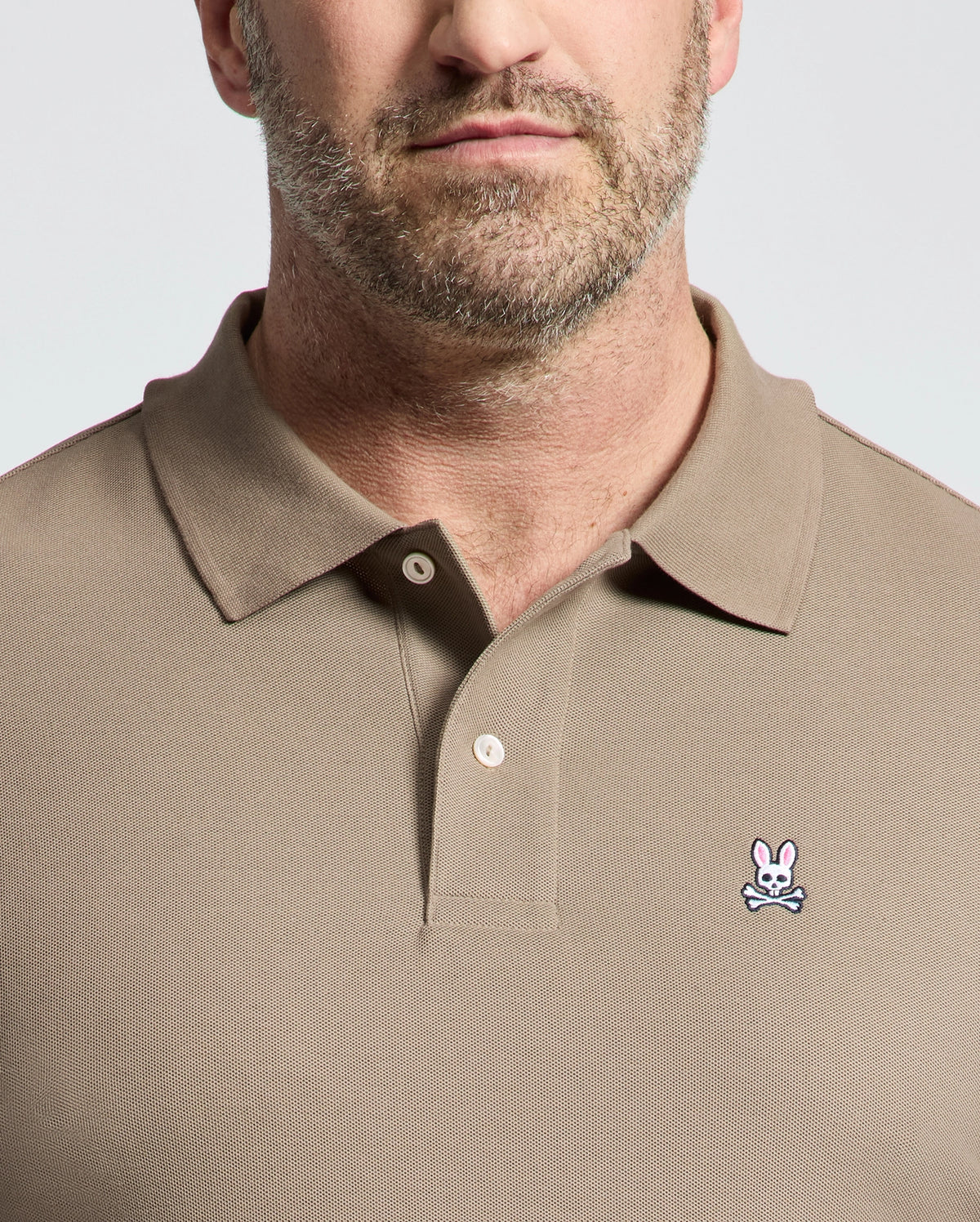 A bearded man wears a Psycho Bunny Men's Big and Tall Classic Pique Polo Shirt (B9K001CRPC) in tan, crafted from soft pima cotton. The shirt features a pink rabbit with crossed bones logo on the left chest and has a buttoned collar against a plain background.