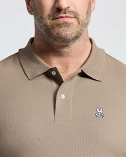 A bearded man wears a Psycho Bunny Men's Big and Tall Classic Pique Polo Shirt (B9K001CRPC) in tan, crafted from soft pima cotton. The shirt features a pink rabbit with crossed bones logo on the left chest and has a buttoned collar against a plain background.