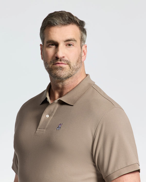 A man with short hair and a beard wears a Psycho Bunny taupe pique polo, crafted from soft pima cotton, featuring an embroidered logo on the chest. He stands against a plain background, looking directly at the camera.