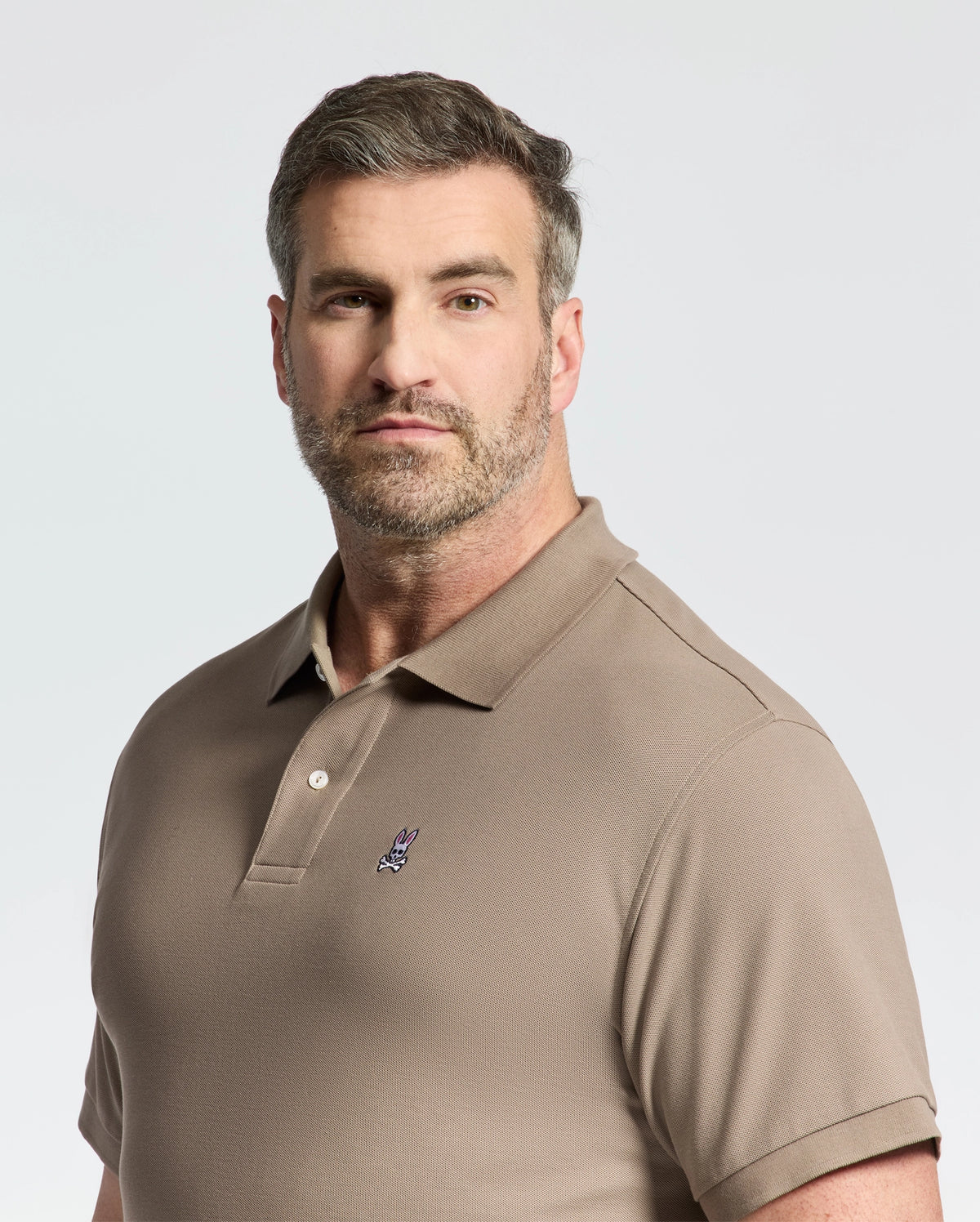 A man with short hair and a beard wears a Psycho Bunny taupe pique polo, crafted from soft pima cotton, featuring an embroidered logo on the chest. He stands against a plain background, looking directly at the camera.