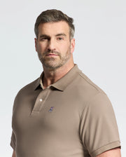 A man with short hair and a beard wears a Psycho Bunny taupe pique polo, crafted from soft pima cotton, featuring an embroidered logo on the chest. He stands against a plain background, looking directly at the camera.
